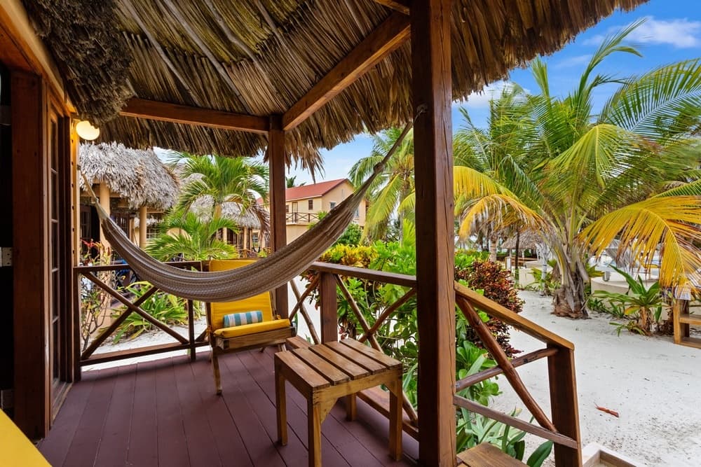 An inviting outdoor patio with a thatched roof, a white hammock, and a wooden table and chair, all surrounded by lush palm trees.