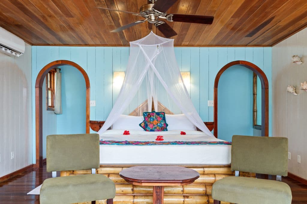 A tranquil bedroom with a king bed draped in a mosquito net, two chairs, and a table. The room features light blue walls, a wooden ceiling, and dark wooden floors.