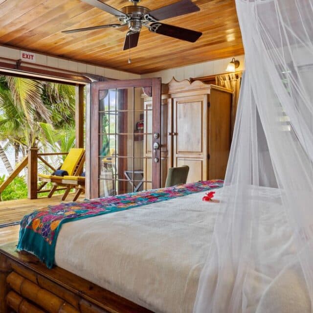 An inviting bedroom with a king bed draped in a mosquito net, leading to an outdoor balcony with lush greenery. The room has wooden ceilings and doors.