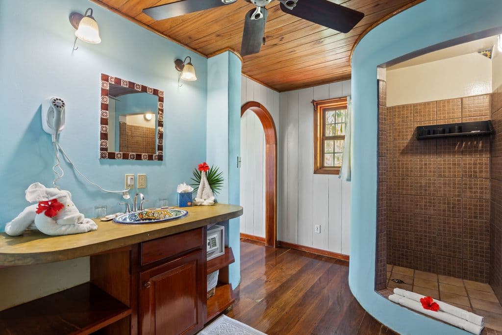 A bathroom with a vanity area, including a mirror, and a walk-in shower. The room has light blue walls, wooden floors, and a wooden ceiling with a fan.