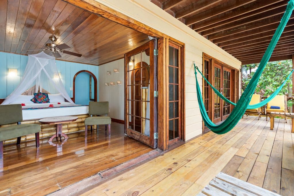 A bedroom with a king bed and a mosquito net, leading to an outdoor patio with two green hammocks and wooden floors.