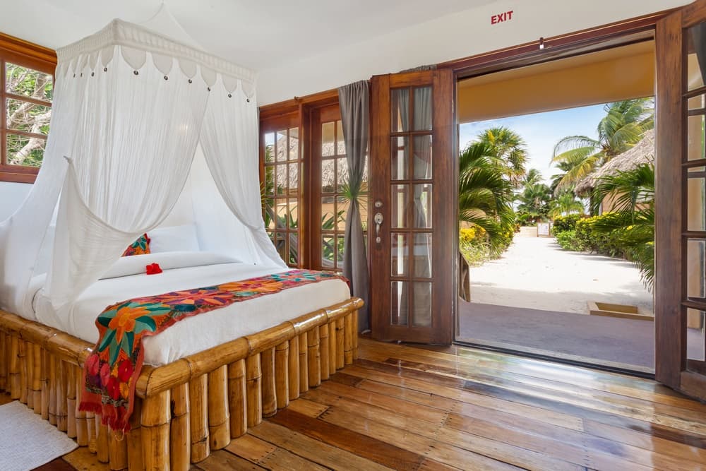 A bright bedroom with a queen bed draped in a white mosquito net, a colorful blanket, and wooden floors, opening to an outdoor path with lush trees.