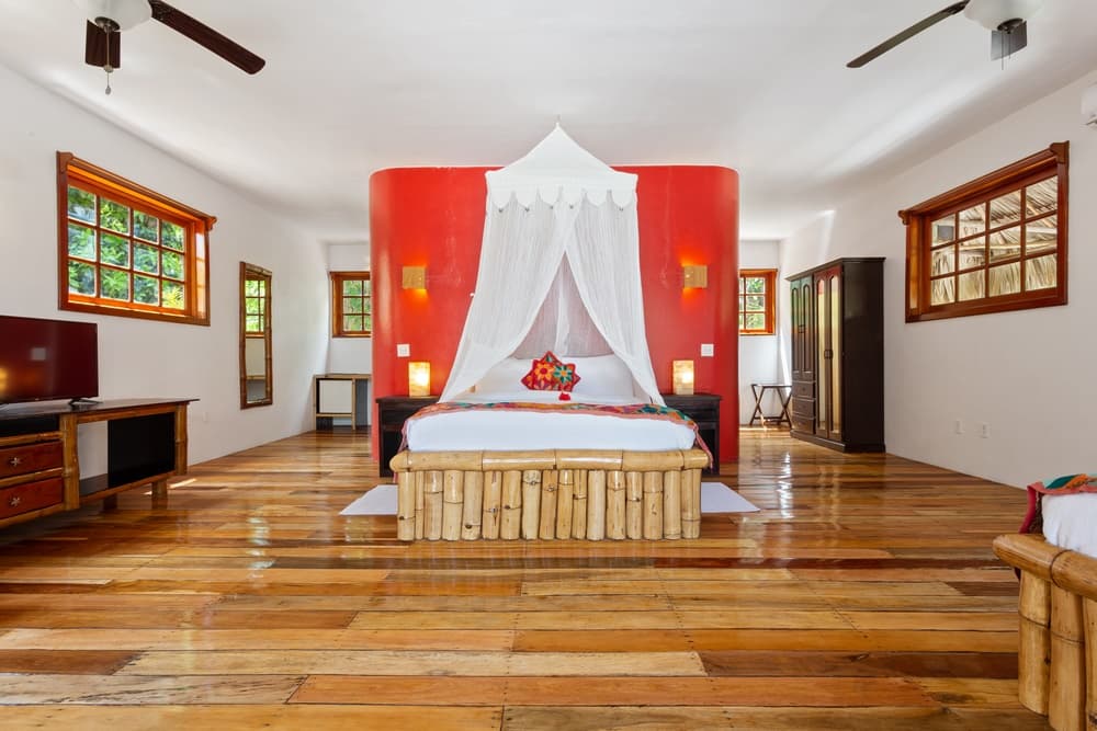 A spacious bedroom with a queen bed featuring a mosquito net and a red headboard wall. The room has wooden floors, multiple windows, and two ceiling fans.