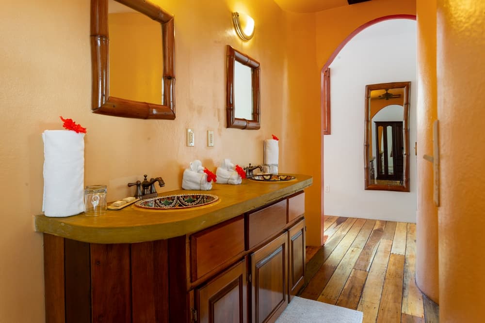 A warm-toned bathroom with a double vanity featuring a dark wooden cabinet and stone sinks. Two mirrors hang above, and an arched doorway leads to another room.