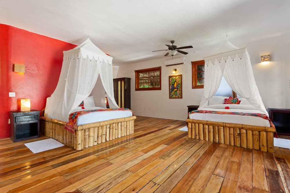 A spacious bedroom with two queen beds, each adorned with a white mosquito net. The room features a red accent wall, wooden floors, and a ceiling fan.