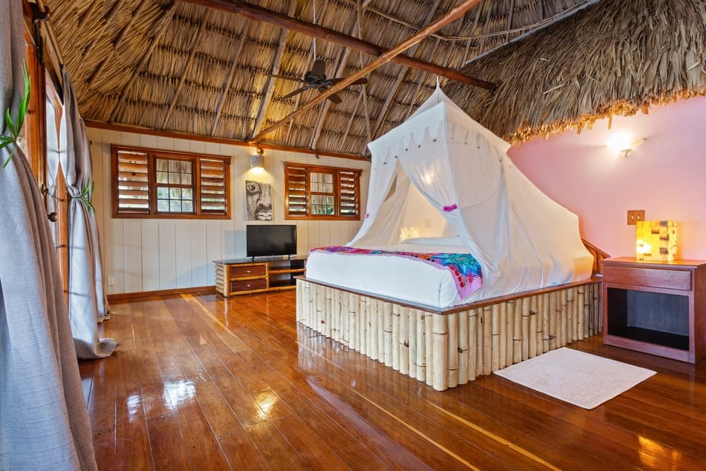 A bedroom with a king bed featuring a white mosquito net and a bamboo frame. The room has a thatched roof, wooden floors, and a TV stand.