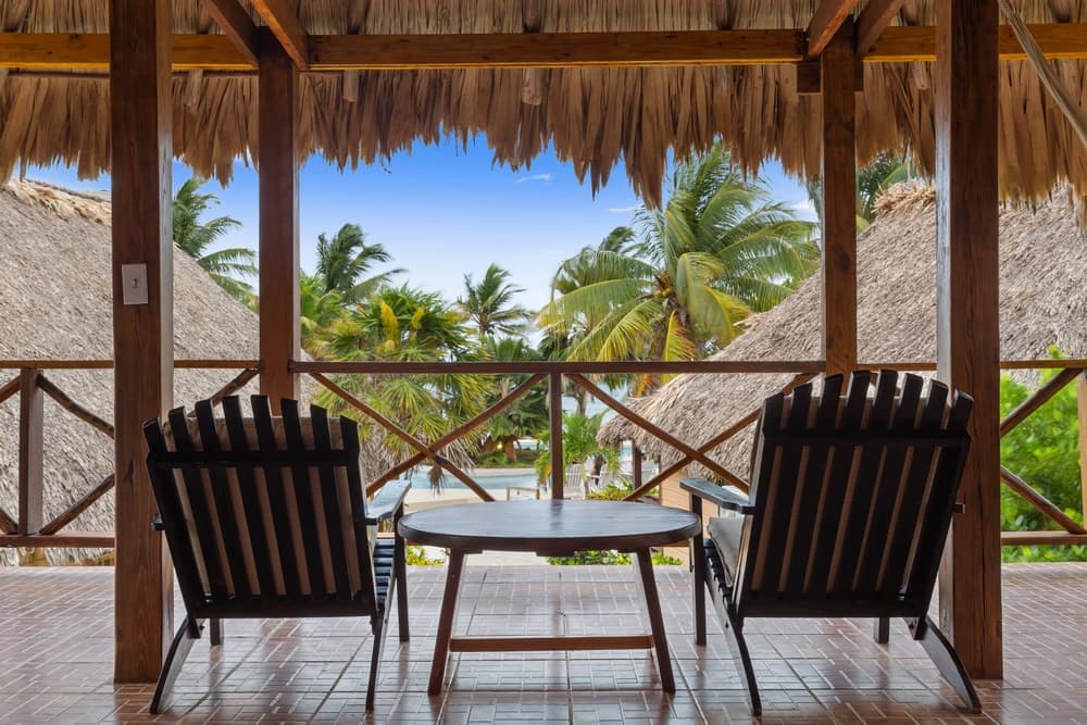 An inviting outdoor patio with a thatched roof, featuring two wooden chairs and a small table, overlooking a tropical landscape with palm trees and blue skies.