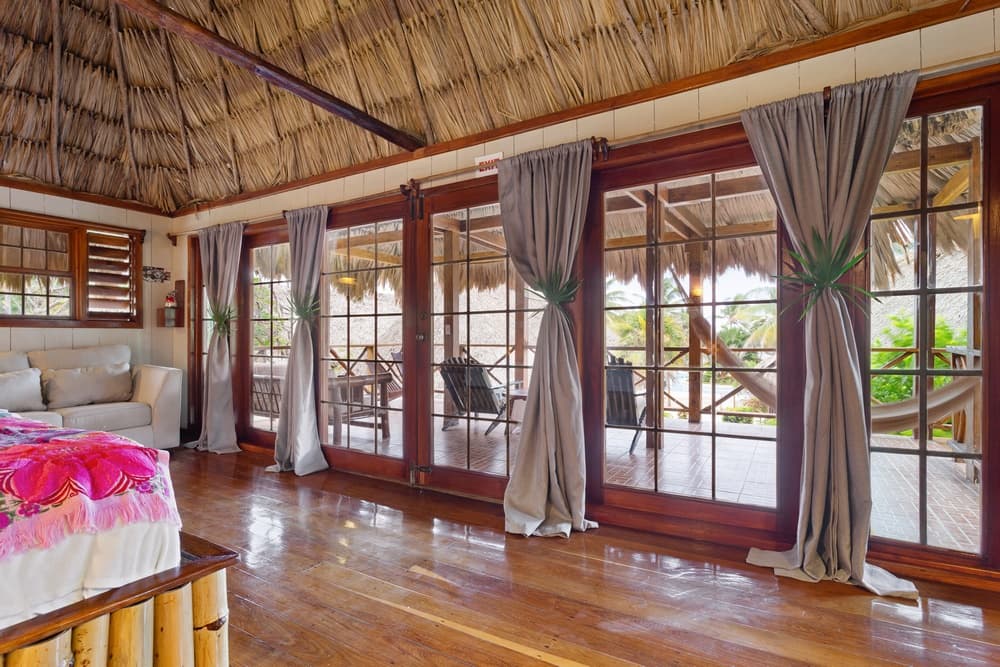 A bedroom room with a thatched roof, wooden floors, and large glass doors with grey curtains, opening to an outdoor patio.