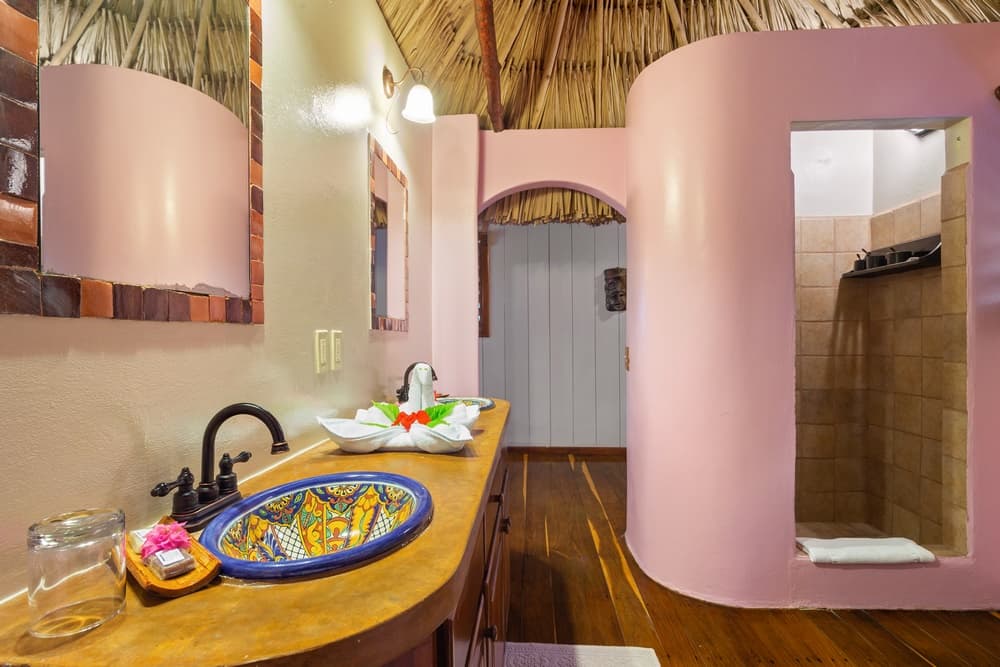 A unique bathroom with a colorful sink, a wooden vanity, a walk-in shower with curved pink walls, and a thatched roof.