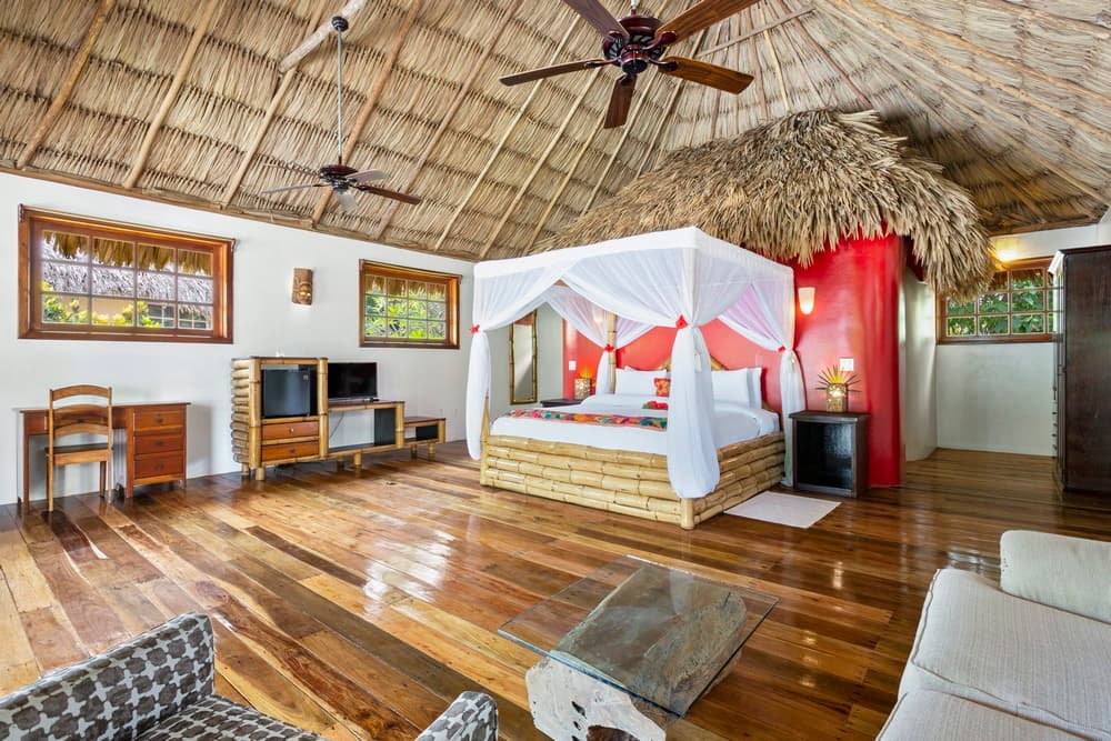 A spacious bedroom with a prominent king bed draped in a white mosquito net, a vibrant red accent wall, and a rustic thatched roof. The room also features wooden floors, a desk, and a seating area.