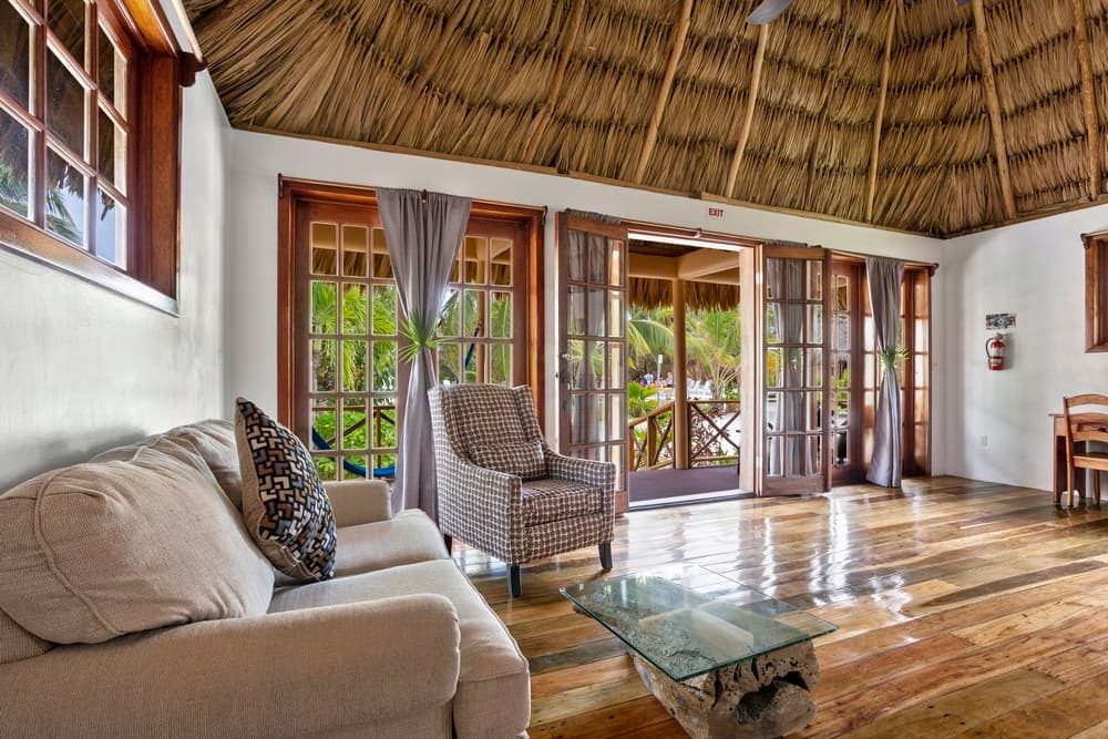 A living room with a thatched roof, wooden floors, and a sofa and armchair. Large glass doors open to an outdoor area with lush greenery.