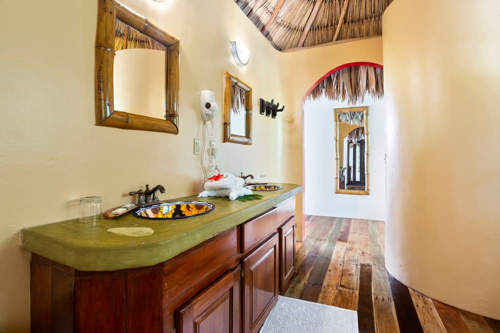 A bathroom with a double vanity featuring a green countertop and wooden cabinets. The room has light yellow walls, wooden floors, and a thatched roof.