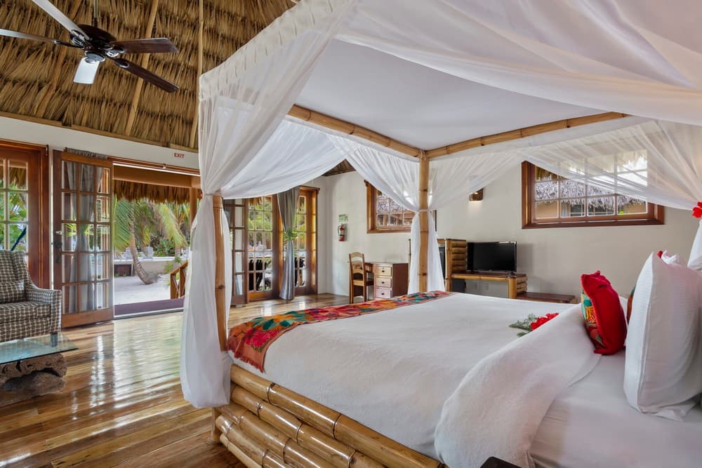 A bedroom with a king bed featuring a white mosquito net and a bamboo frame. The room has a thatched roof, wooden floors, and opens to an outdoor area.