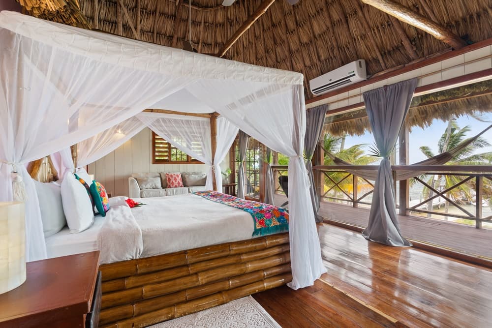 A bedroom with a king bed featuring a white mosquito net and a bamboo frame. The room has a thatched roof, wooden floors, and large windows overlooking palm trees.