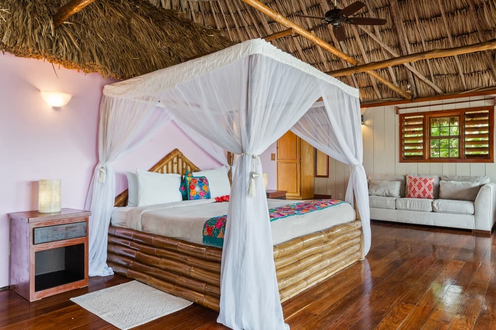A spacious bedroom with a king bed featuring a white mosquito net and a bamboo frame. The room has a thatched roof, wooden floors, and a seating area.