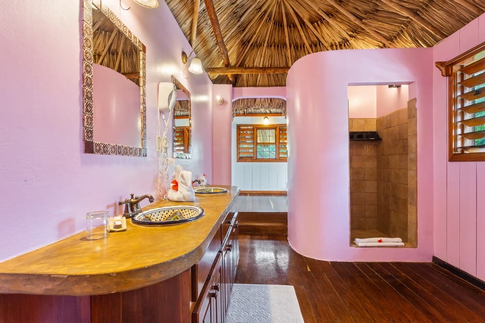 A vibrant bathroom with a pink wall, a long wooden vanity with two sinks and mirrors, a walk-in shower, and a thatched roof.