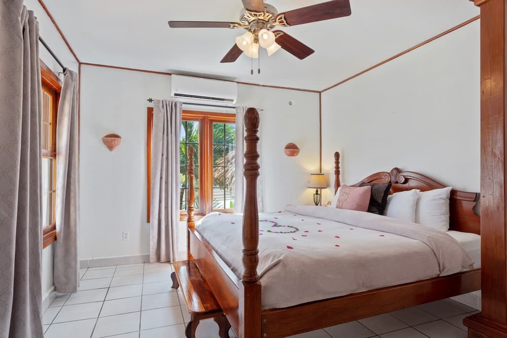 A bedroom with a four-poster wooden bed adorned with rose petals. The room has white walls, tiled floors, a ceiling fan, and access to an outdoor area.