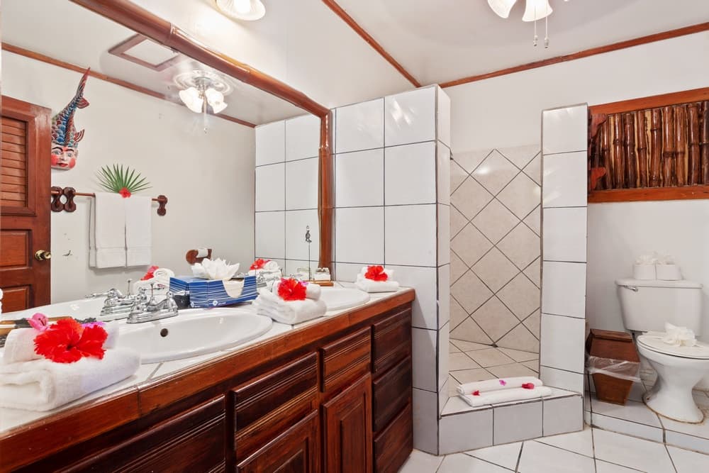 A bright bathroom with a long wooden vanity featuring a double sink. The room has white tiled walls, a walk-in shower, and a toilet area.