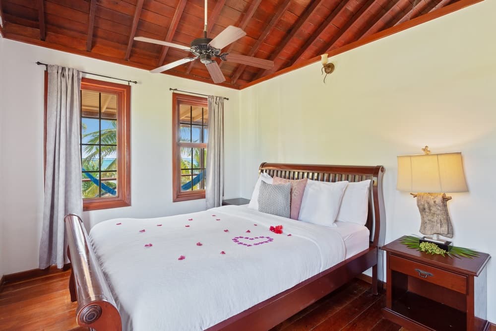 A bedroom with a wooden bed adorned with rose petals. The room features a wooden ceiling, ceiling fan, and windows offering an outdoor view.