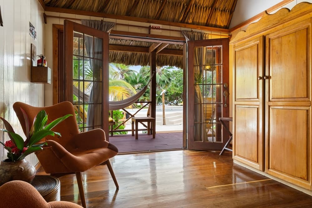 An indoor living area with two orange armchairs and wooden cabinets, opening up to an outdoor patio with a hammock and lush greenery.