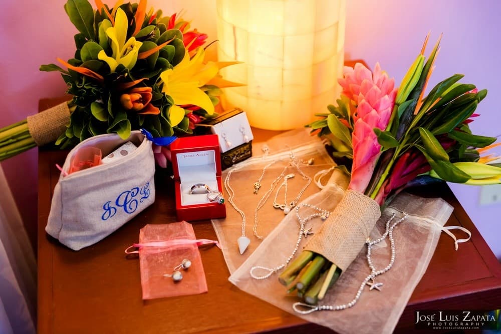 A collection of jewelry, a bouquet of flowers, and a cosmetic bag arranged on a wooden table near a lamp.