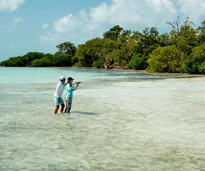Two anglers stand in shallow water, casting lines toward a shoreline of greenery under a blue sky.