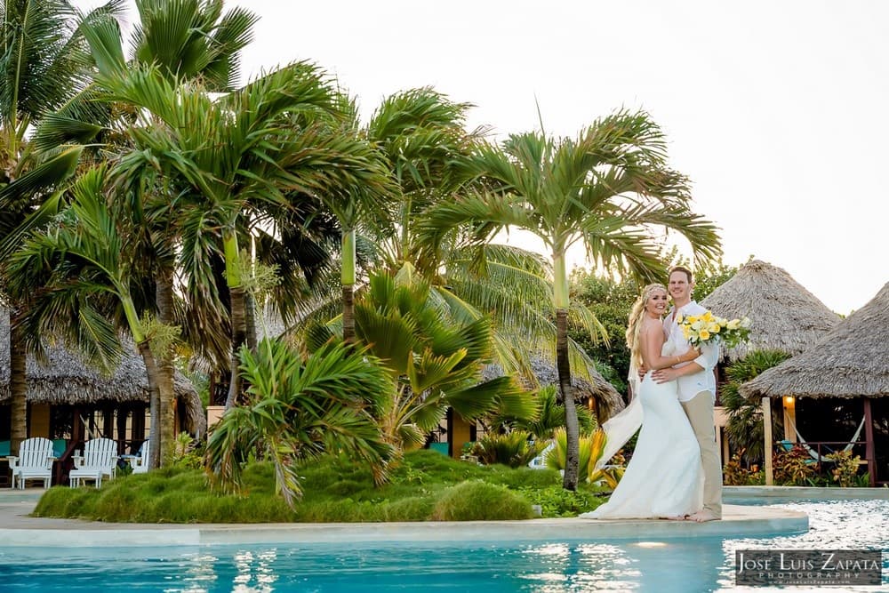 A smiling couple in wedding attire stands by a tropical pool, surrounded by palm trees.