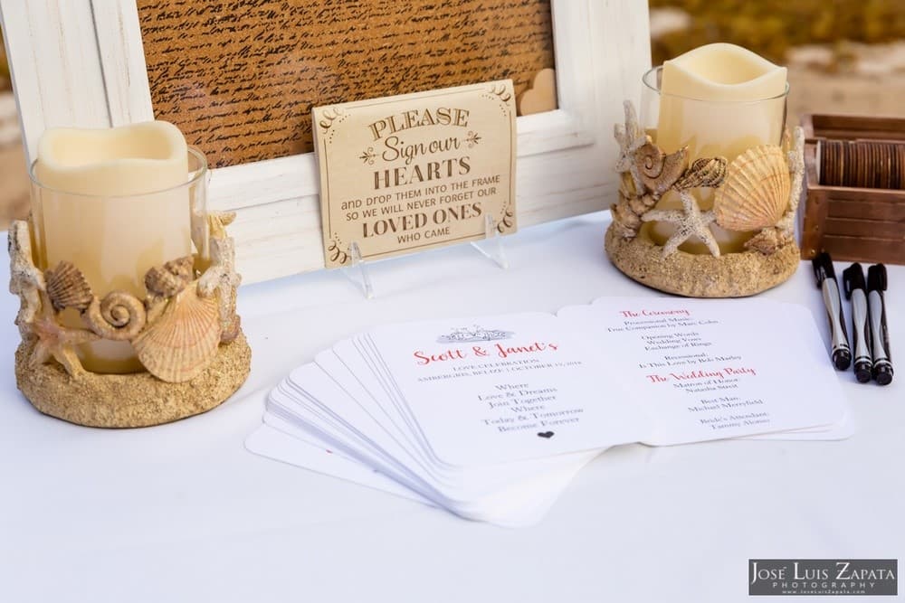 A wedding display featuring a sign for guests to sign hearts, candles in sandy shells, and an event program.
