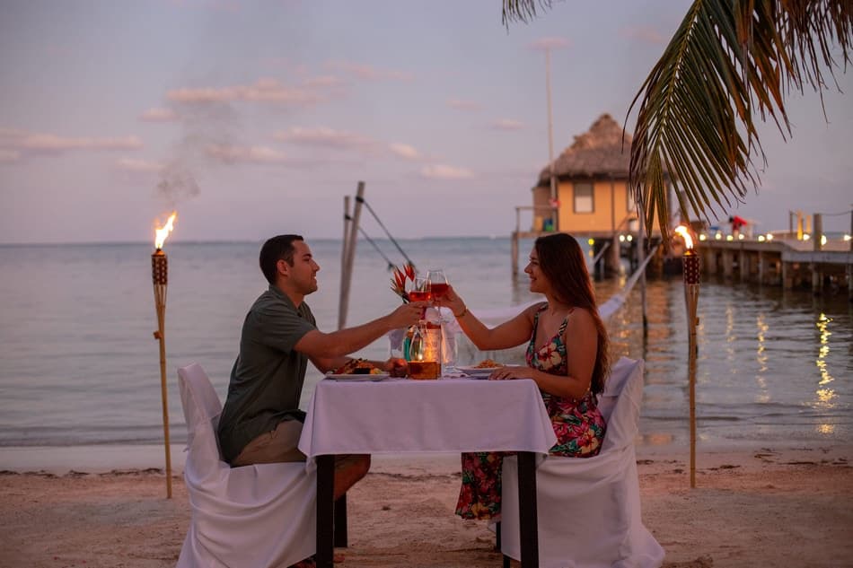 A couple clinks glasses during a romantic beachside dinner at sunset.