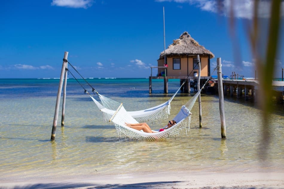 A person relaxes on a hammock by a tranquil beach with a thatched-roof hut in the background.