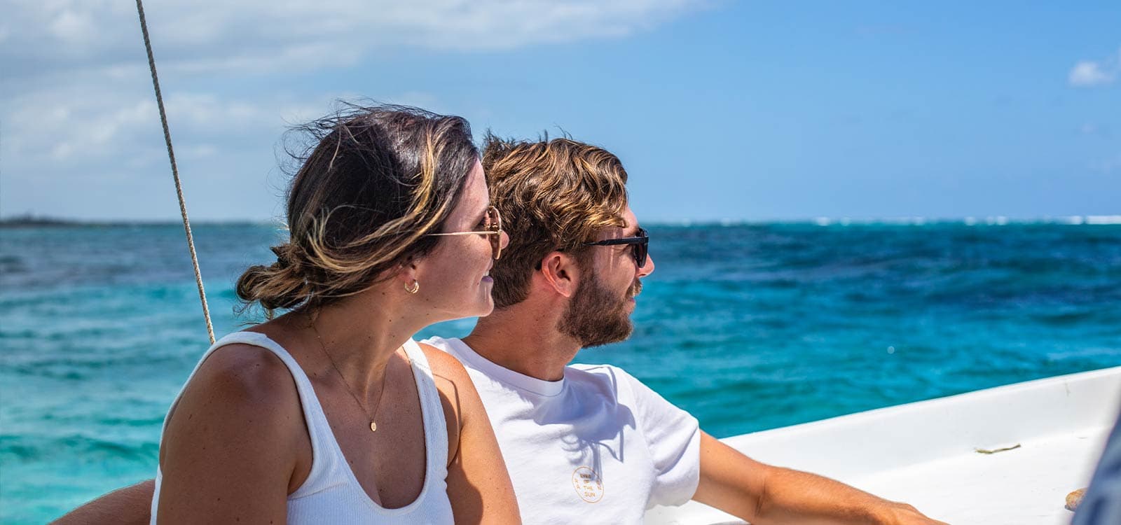 A couple wearing sunglasses looks out at the ocean while sitting on a boat. The sky is clear, and the water is vibrant blue, creating a serene atmosphere.