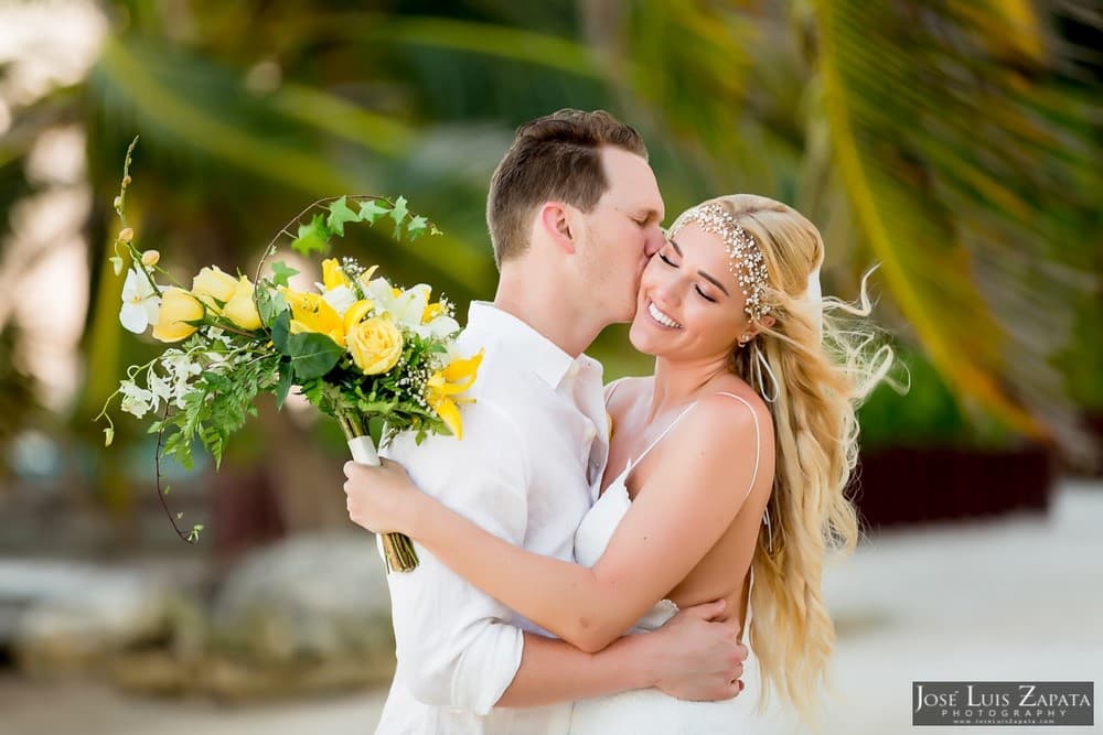 A couple joyfully embraces, surrounded by a tropical backdrop, with the woman holding a bouquet of yellow and white flowers.