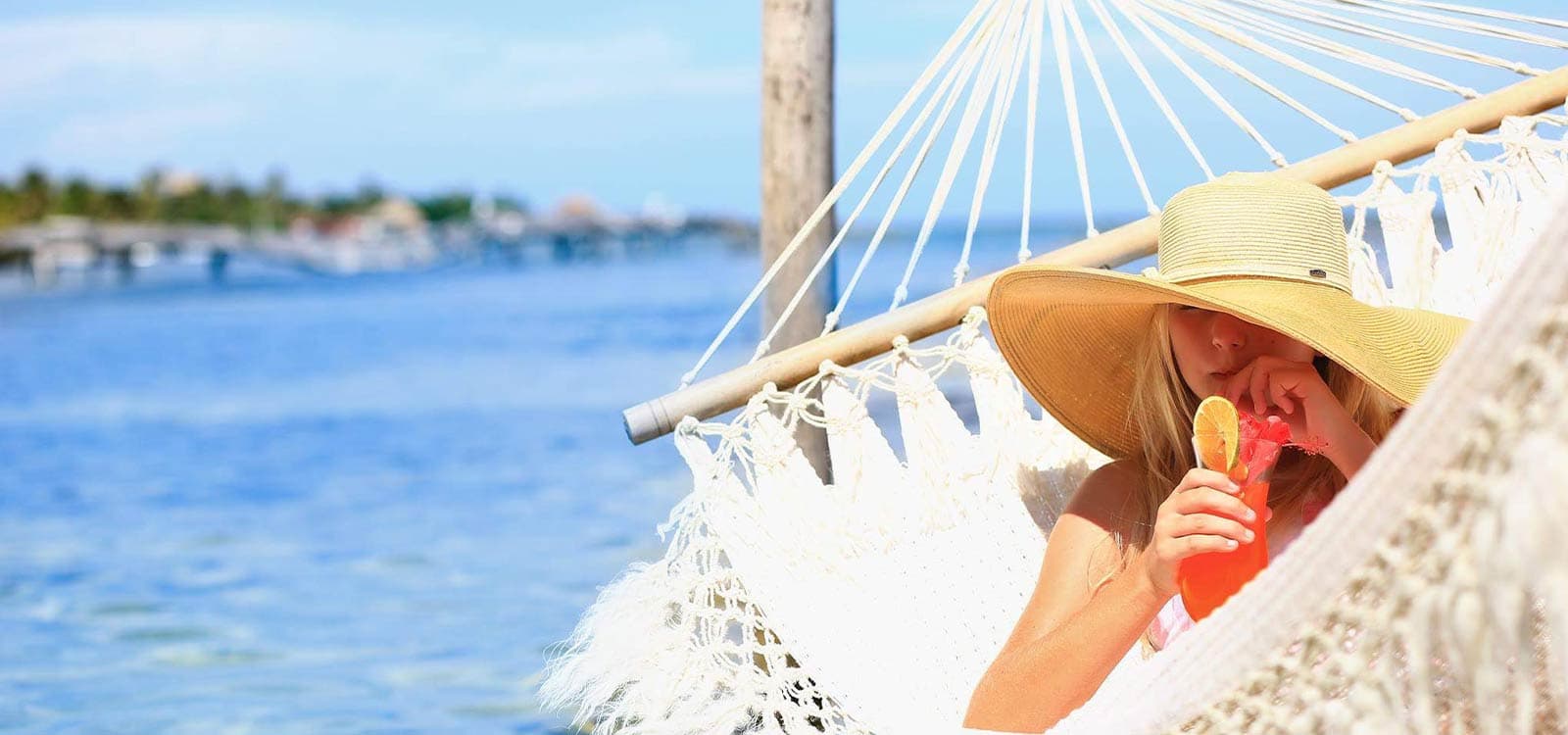 A woman with a wide-brimmed hat lounges in a white crocheted hammock on a beach, while sippoing a frozen red cocktail