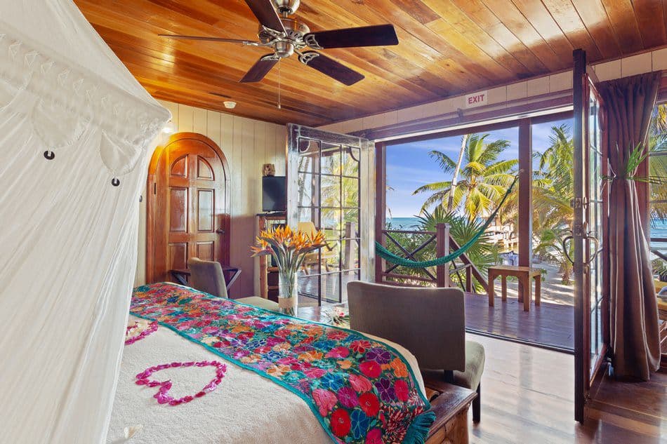 A cozy room with a floral bedspread, a hammock, and a beachfront view through large open windows.