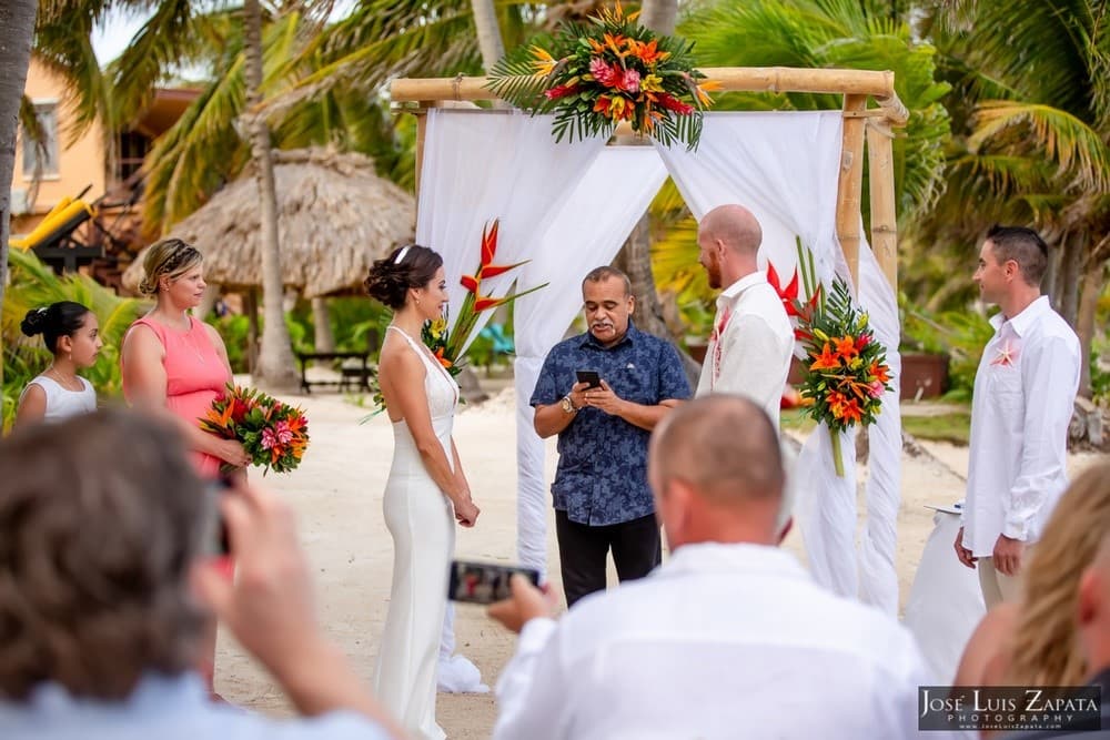A couple stands under a floral arch during their wedding ceremony on a beach, surrounded by guests.