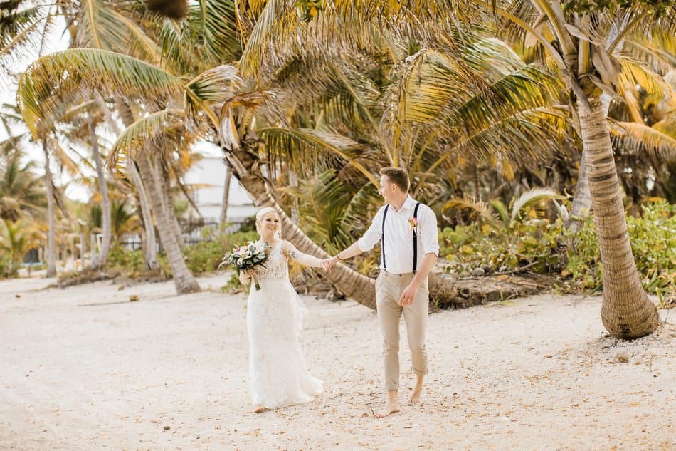 A newlywed couple holds hands while walking along a sandy beach adorned with palm trees.