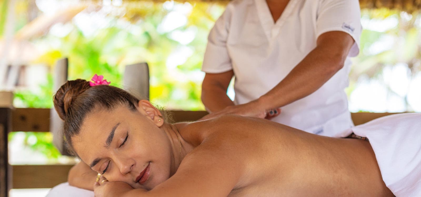 Woman receiving a relaxing back massage outdoors under a thatched roof. She's lying on a massage table, with eyes closed and a peaceful expression.