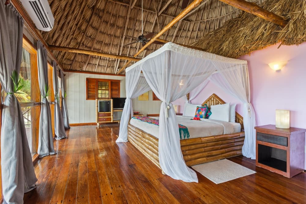 A bedroom with a king bed featuring a white mosquito net and a bamboo frame. The room has a thatched roof, wooden floors, and a pink accent wall.
