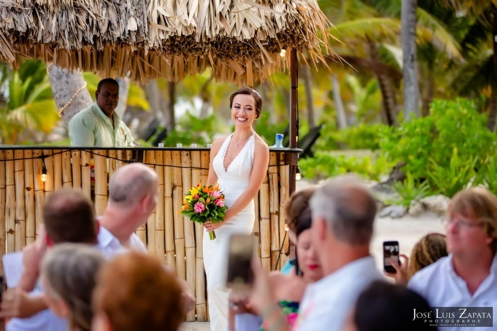 A bride smiles while holding a bouquet, with guests looking on at a tropical wedding ceremony.