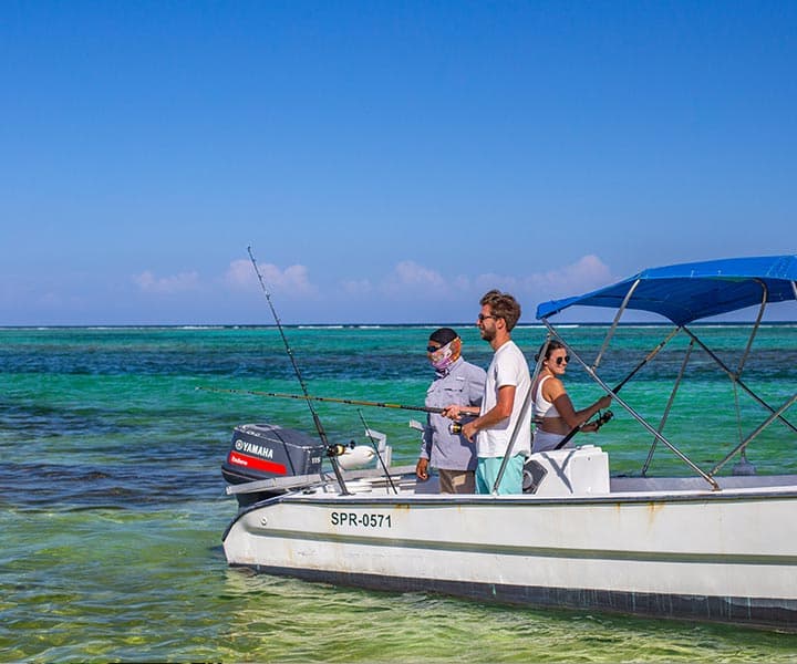 Three people are fishing on a small boat in clear, turquoise waters under a bright blue sky. They appear relaxed, enjoying a sunny day.
