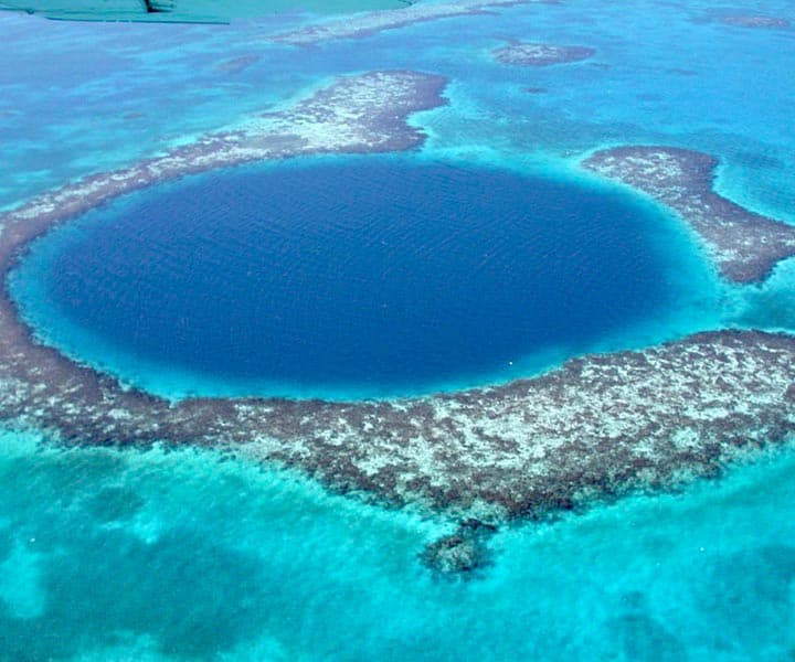 Aerial view of a circular blue hole surrounded by turquoise water and coral reefs.