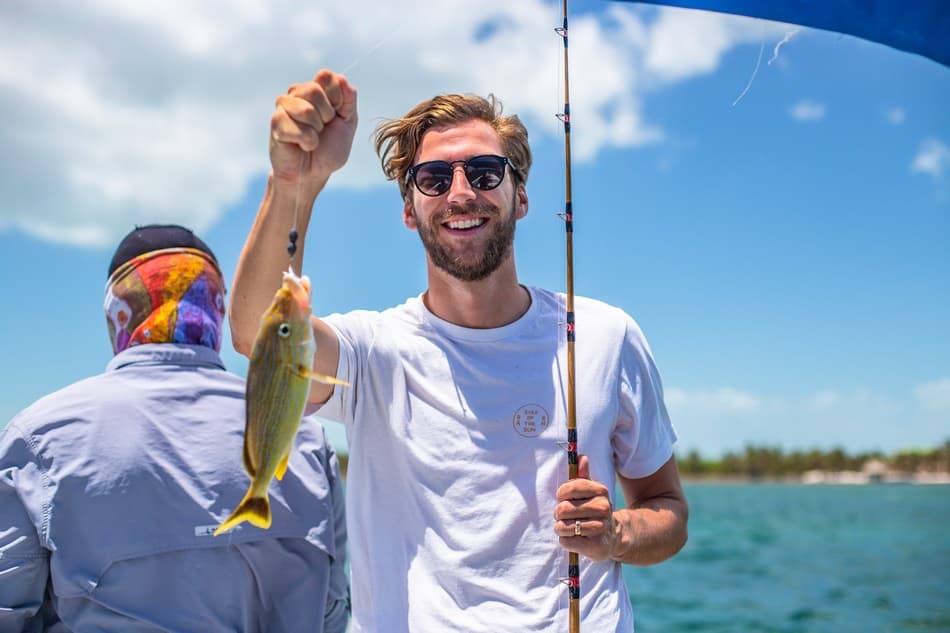 A smiling man in sunglasses holds a fishing rod with a caught fish against a sunny, blue sky backdrop.
