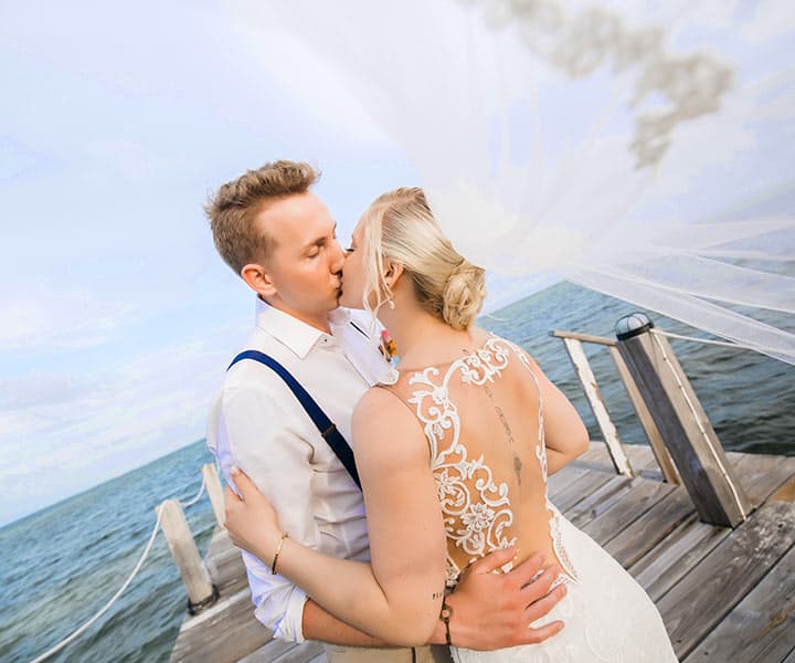 A bride and groom kiss on a dock by the water, with the bride's veil gently flowing in the breeze.