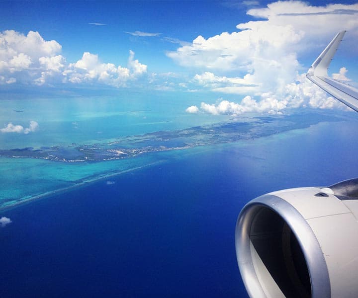 View from airplane window showing airplane wing and engine above vibrant blue ocean and green land, under a vast sky with fluffy clouds. Calm and serene.