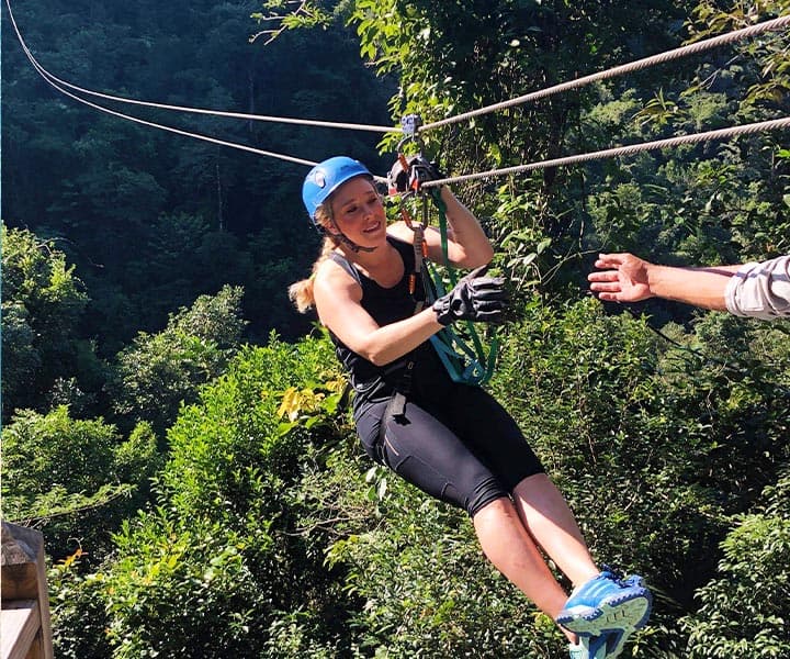A person zip-lining through a lush green landscape, reaching for a hand from a guide.