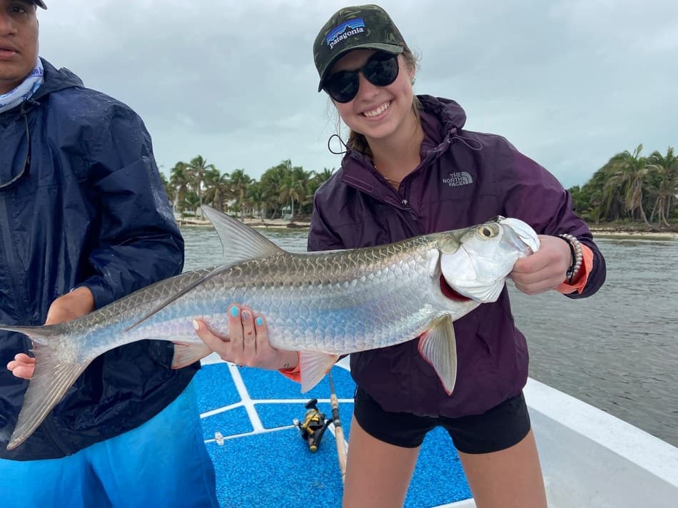 A woman smiles while holding a large fish on a boat, with a tropical landscape in the background.