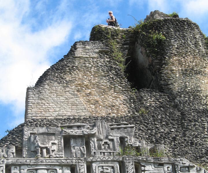 A person sits atop a stone pyramid surrounded by lush greenery under a blue sky.